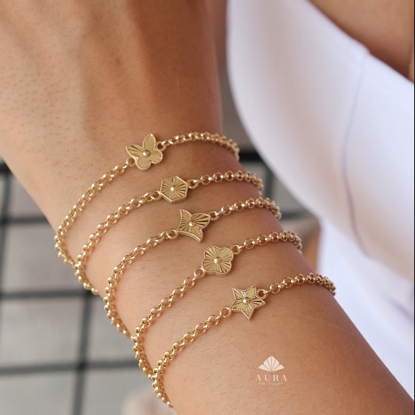 May include: Gold-toned charm bracelets with various designs, including a butterfly, hexagon, flower, clover, and star. The bracelets are displayed on a wrist, showcasing the delicate chain links and detailed charms. The jewelry is elegant and suitable for everyday wear.