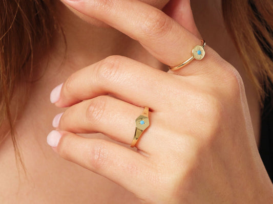May include: Gold rings with turquoise starburst designs. One ring features a round face, while the other has a hexagonal face. The rings are worn on a hand with a light skin tone.