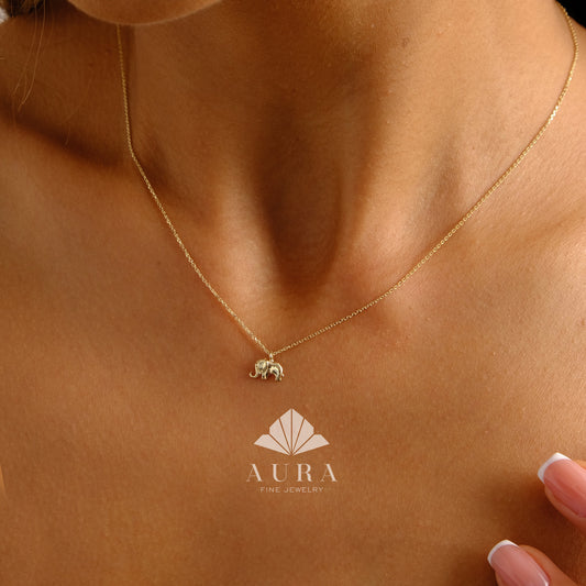 May include: A gold chain necklace with a small elephant pendant. The necklace is on a model's neck. The brand name "AURA FINE JEWELRY" is visible at the bottom of the image.
