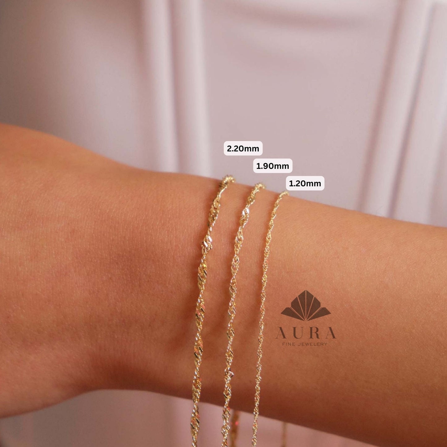 May include: Three gold chains of different thicknesses, 2.20mm, 1.90mm, and 1.20mm, are shown on a person's wrist. The chains are made of a delicate, twisted design. The image includes the text "AURA FINE JEWELRY".