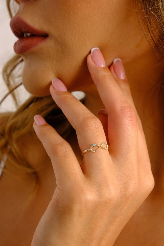 May include: A gold infinity ring with a blue evil eye charm. The ring is worn on a person's finger.