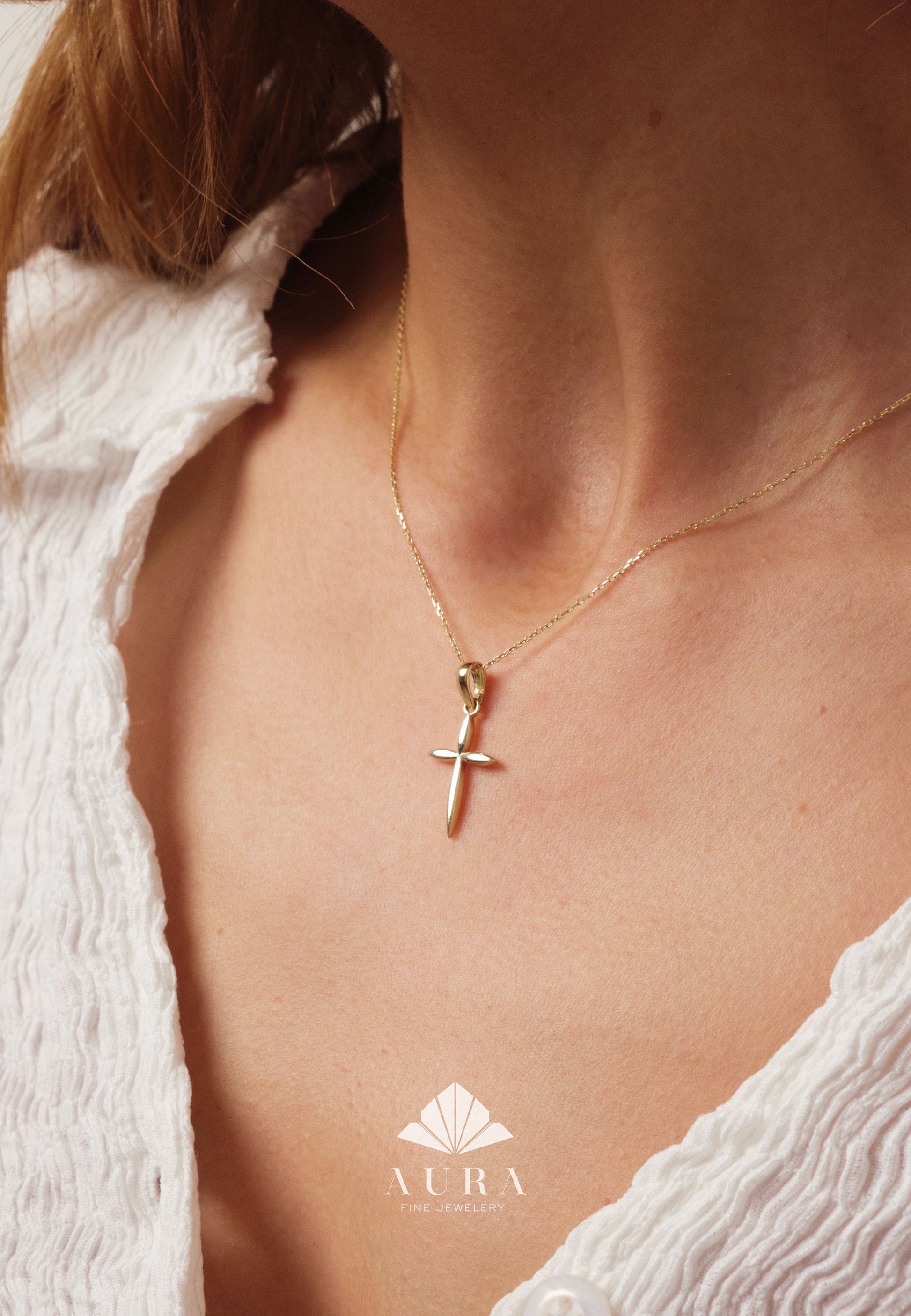 May include: A gold chain necklace with a small, simple gold cross pendant. The necklace is worn by a person with a white shirt.