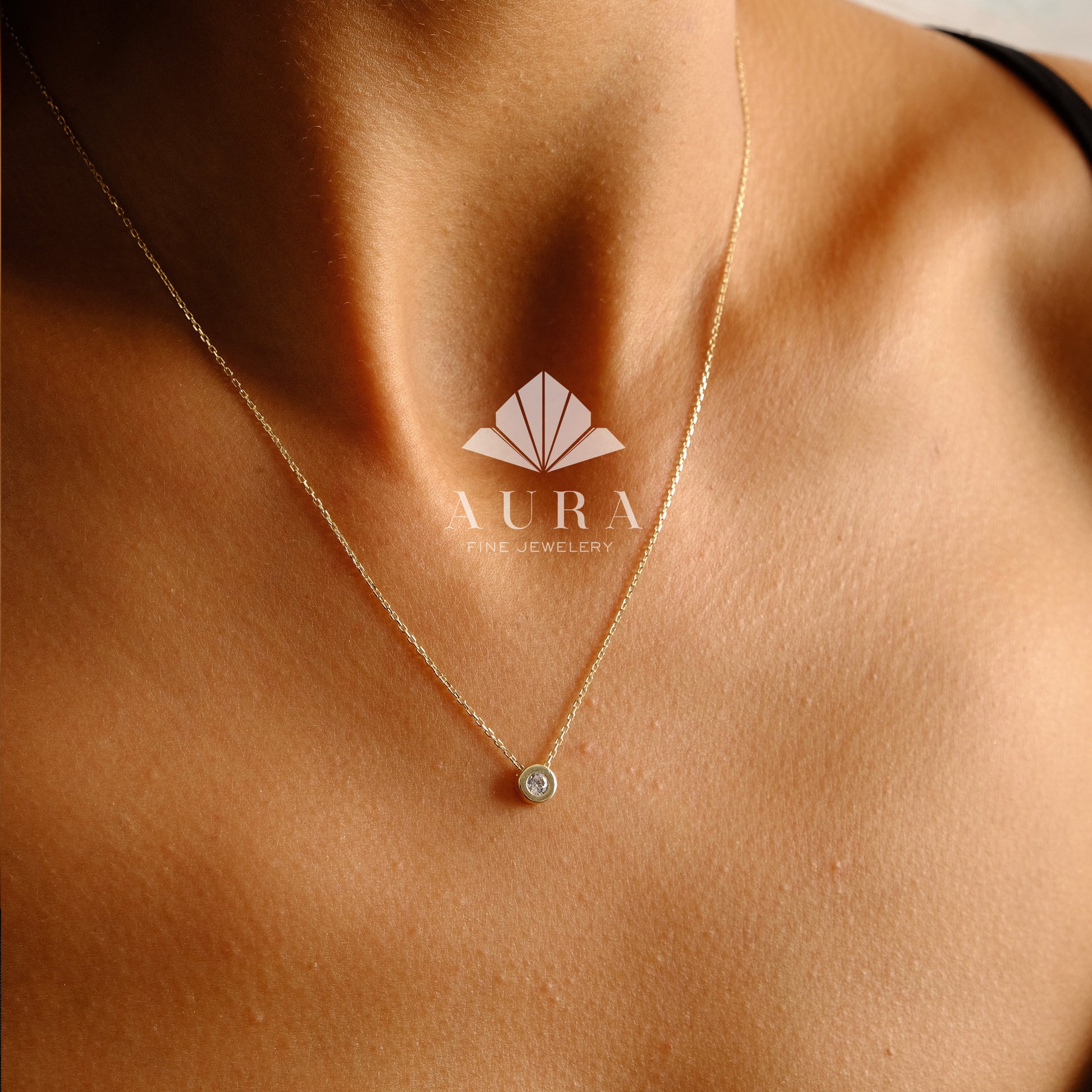 May include: A delicate gold chain necklace with a small, round, clear gemstone pendant. The necklace is worn by a person with a tan complexion. The text "AURA FINE JEWELRY" is visible in the image.