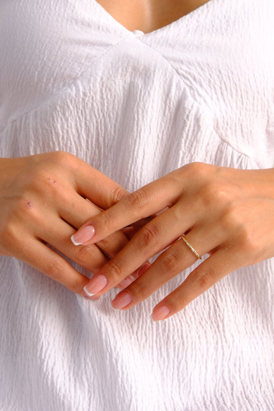 May include: A close-up of a hand wearing a thin gold band ring. The ring is twisted and has a polished finish. The hand is resting on a white fabric surface.